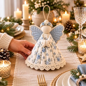 Pu&ograve; includere: Un ornamento a forma di angelo in tessuto con testa color crema, corona di fiori blu, ali trapuntate azzurre e un abito conico con motivo floreale blu su sfondo crema, rifinito con un bordo in pizzo all'uncinetto bianco alla base.