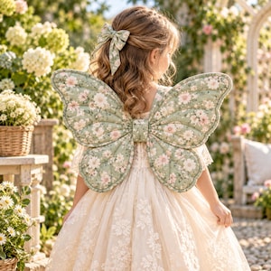 May include: A pair of quilted butterfly wings in a sage green fabric with a pink and white floral pattern, worn over a cream-colored dress with lace details. A matching sage green floral hair bow is pinned to the back of the hair.