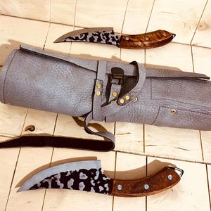 Könnte beinhalten: Zwei gebogene Messer mit dunkel gemusterten Klingen und braunen Holzgriffen, begleitet von einer hellgrauen, strukturierten Lederrolltasche mit Metallschnallenverschluss und Messingnieten, angeordnet auf einer hellen Holzoberfläche.