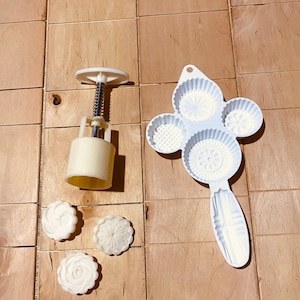Op de afbeelding: Een wit plastic veerbelast stempelgereedschap voor het vormen van deeg, vergezeld van een witte plastic vormplaat met vier verschillende cirkelvormige patronen en drie kleine, ronde, witte gevormde deegstukjes met bloem- en geometrische ontwerpen.