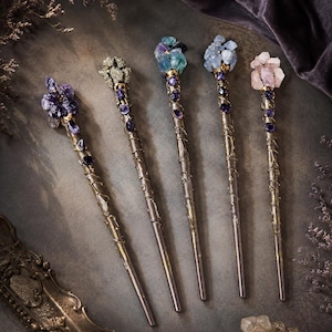 May include: Five metal hair sticks featuring decorative tops adorned with clusters of raw crystals and stones in purple, metallic gold, blue, light blue, and pink, with additional small dark purple beads wrapped along the length of each metal shaft.