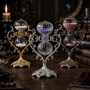 May include: Three ornate hourglasses with glass bulbs and metal frames. The left one is gold with white sand, the center is silver with purple sand and floral accents, and the right one is bronze with pink sand. Each features a heart-shaped frame.