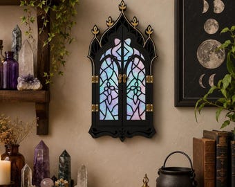 May include: A black wall-mounted cabinet with a pointed gothic arch shape, featuring two doors with intricate cut-out patterns backed by iridescent, multi-colored panels. The cabinet includes gold-toned metal hinges and a central latch closure.