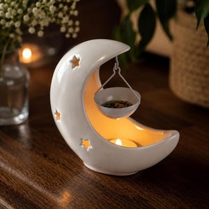 May include: A white ceramic crescent moon oil burner featuring star-shaped cutouts. A small white metal bowl hangs from the top of the moon, and a tea light candle sits in the base, illuminating the interior of the moon-shaped structure.