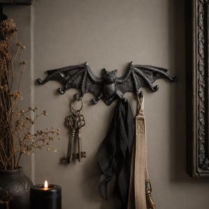 May include: A black, cast-metal wall hook shaped like a bat with outstretched wings. The bat features multiple hooks along its lower edge, currently holding a set of vintage-style metal keys, a black fabric item, and a beige strap with a metal clip.