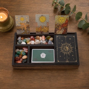 May include: A dark wooden organizer tray containing three upright tarot cards, a deck of cards with a green back, a black card box with gold celestial designs, and two compartments filled with assorted polished multi-colored gemstones.