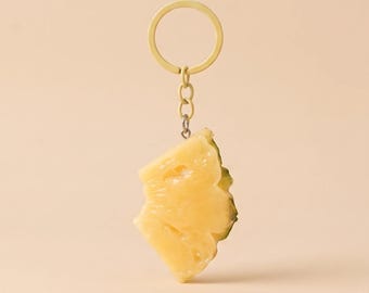 May include: A keychain featuring a yellow pineapple slice charm attached to a gold-toned metal ring. The text PINEAPPLE SLICE CHARM KEYCHAIN and Delicate & Handmade is printed above the charm. The charm has a textured, translucent appearance.