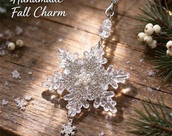Könnte beinhalten: Ein transparenter Acryl-Schneeflocken-Anhänger mit einer zentralen Anordnung aus weißen Perlen, silberfarbenen Strass-Akzenten und einem kleinen, baumelnden Schneeflocken-Anhänger. Mit silberfarbenem Metallverschluss und weißer Kordel.