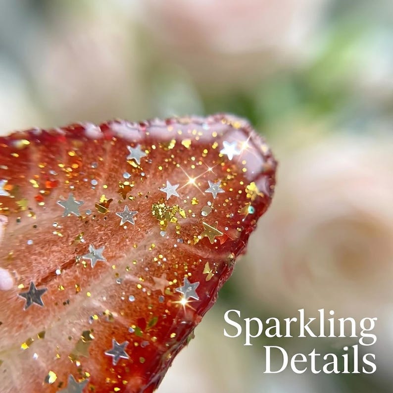 May include: A translucent, reddish-orange resin object shaped like a strawberry slice, embedded with numerous small silver star-shaped sequins and fine gold glitter particles throughout the surface, featuring the text Sparkling Details.