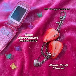 K&ouml;nnte beinhalten: Ein rosa Klapphandy und ein silberner Kettenanh&auml;nger mit zwei Erdbeerscheiben, die mit Metallpiercings, einer Sicherheitsnadel und einem kleinen Herzanh&auml;nger verziert sind. Kleine durchsichtige Kunststoffschmetterlinge liegen verstreut dabei.