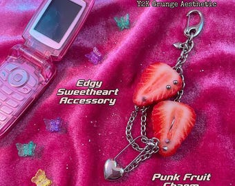 Könnte beinhalten: Ein rosa Klapphandy und ein silberner Kettenanhänger mit zwei Erdbeerscheiben, die mit Metallpiercings, einer Sicherheitsnadel und einem kleinen Herzanhänger verziert sind. Kleine durchsichtige Kunststoffschmetterlinge liegen verstreut dabei.