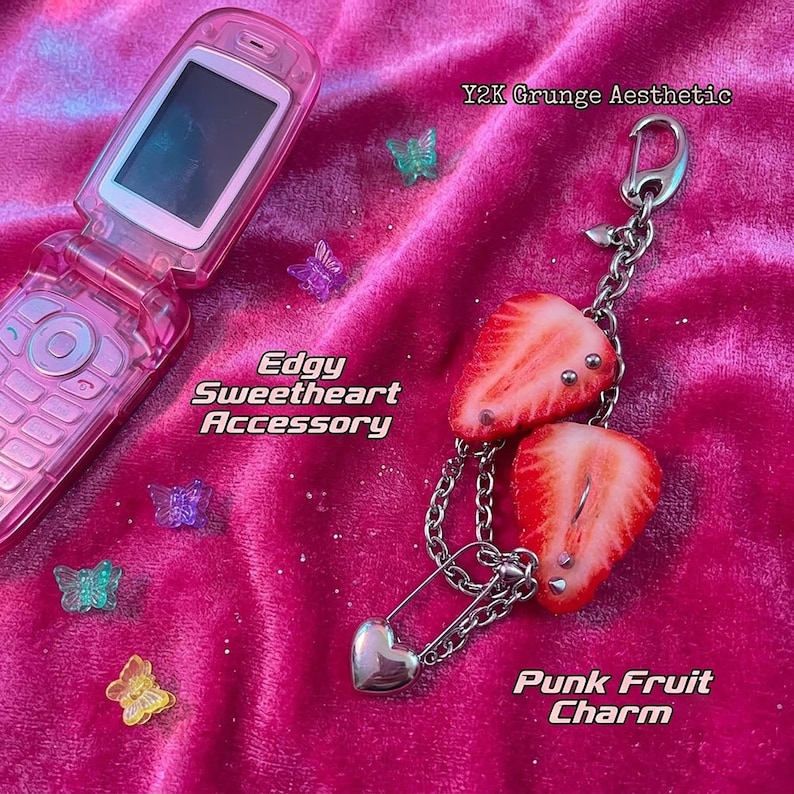 K&ouml;nnte beinhalten: Ein rosa Klapphandy und ein silberner Kettenanh&auml;nger mit zwei Erdbeerscheiben, die mit Metallpiercings, einer Sicherheitsnadel und einem kleinen Herzanh&auml;nger verziert sind. Kleine durchsichtige Kunststoffschmetterlinge liegen verstreut dabei.
