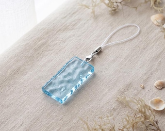 Könnte beinhalten: Ein rechteckiger, hellblauer Glasanhänger mit einer wellenförmigen, wasserartigen Textur. Der Anhänger ist an einem silberfarbenen Metallring und einer dünnen weißen Kordelschlaufe befestigt, die als Befestigung für Handys oder Zubehör dient.