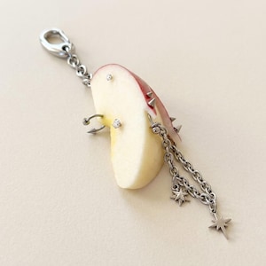 May include: A silver-toned metal charm featuring a slice of red apple adorned with multiple silver piercings, including small studs, a hoop, and spikes. A silver chain hangs from the apple, decorated with two small star-shaped metal charms.