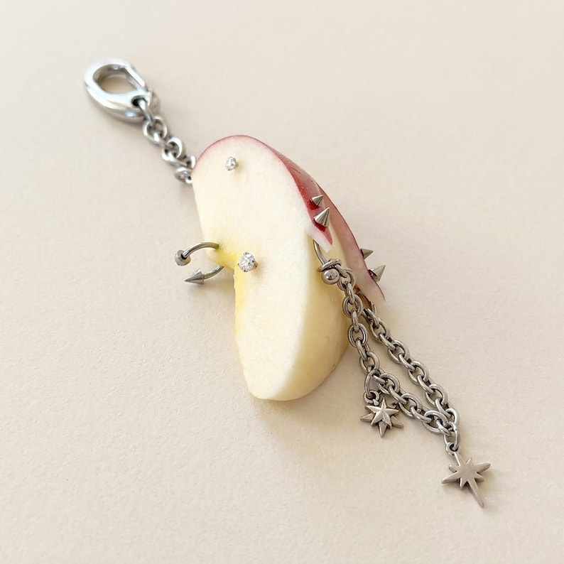 May include: A silver-toned metal charm featuring a slice of red apple adorned with multiple silver piercings, including small studs, a hoop, and spikes. A silver chain hangs from the apple, decorated with two small star-shaped metal charms.