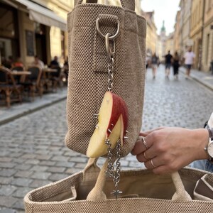 May include: A beige textured fabric tote bag featuring a hanging charm accessory. The charm consists of a red and white apple slice decorated with silver metal spikes and a dangling silver chain with small star-shaped pendants attached to it.