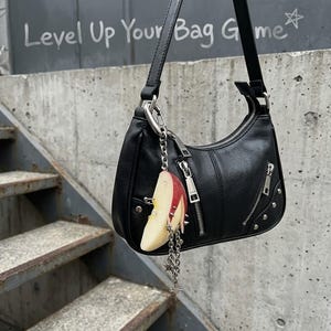May include: A black shoulder bag with a zipper closure, two diagonal front zipper pockets, and silver-tone metal studs. A silver chain charm featuring a realistic apple slice with metal piercings and dangling star charms hangs from the strap.