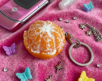 May include: A keychain featuring a realistic orange citrus fruit segment charm attached to a silver metal ring. The scene includes a pink flip phone, a clear lip gloss tube, small colorful butterfly charms, silver star charms, and rhinestones.