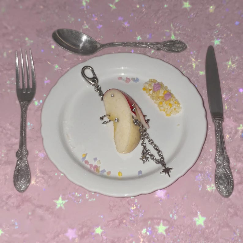 May include: A white ceramic plate holds a sliced apple segment adorned with silver-toned metal piercings, including rings and studs, attached to a silver chain with star-shaped charms. A silver spoon and knife flank the plate on a pink surface.
