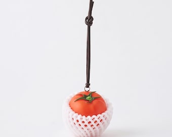 Könnte beinhalten: Eine leuchtend rote Tomate mit grünem Stiel, die in einem weißen Schutznetz aus Schaumstoff steckt. Die Tomate hängt an einem dünnen, dunkelbraunen Lederband mit einem einfachen Knoten am oberen Ende, das als hängendes Dekorationsobjekt dient.
