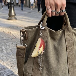 May include: An olive green canvas tote bag featuring a silver-toned metal keychain. The keychain consists of a realistic apple slice charm adorned with small silver spikes, attached to a silver chain with dangling star-shaped charms and a clip.