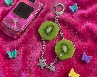 Könnte beinhalten: Ein rosa Motorola-Klapphandy und ein silberner Schlüsselanhänger mit zwei grünen Kiwischeiben, kleinen silbernen Sternanhängern und klaren Strasssteinen, umgeben von mehreren kleinen, bunten, schmetterlingsförmigen Kunststoffteilen.