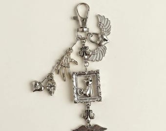 Könnte beinhalten: Ein silberfarbener Metall-Schlüsselanhänger mit Karabinerverschluss, einem geflügelten Herzanhänger, einer Skeletthand, einem kleinen Totenkopf, einem Herzanhänger und einem rechteckigen Rahmenanhänger mit Katzenfigur, ergänzt durch ein geflügeltes Herz.