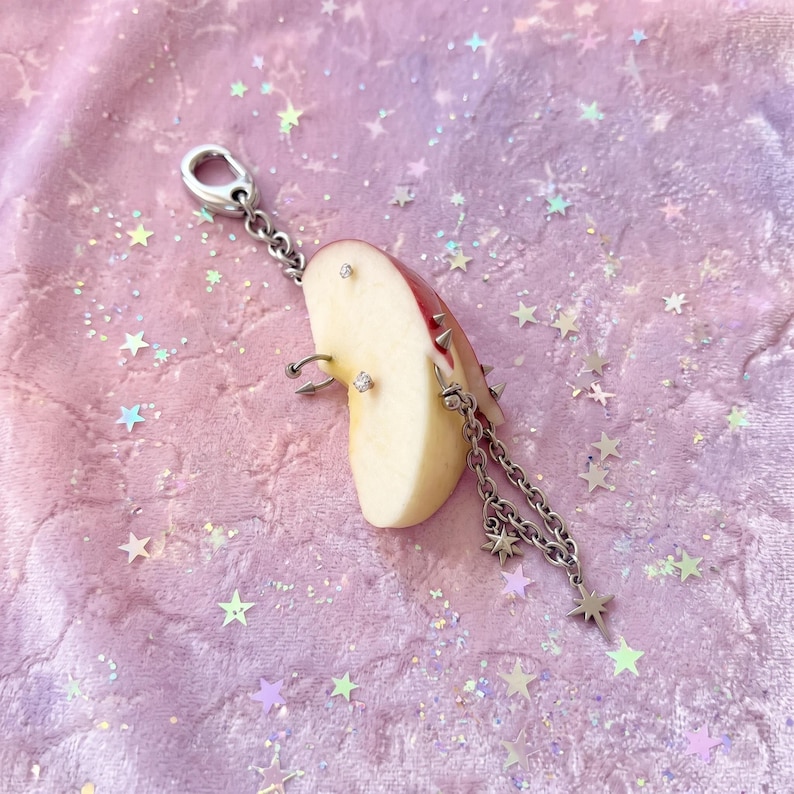 May include: A silver-toned metal keychain featuring a realistic apple slice charm. The apple slice is decorated with multiple silver piercings, including small studs, a hoop, and spikes. A silver chain with star-shaped charms hangs from the apple.