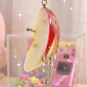 May include: A keychain featuring a realistic apple slice charm with a red skin and pale yellow flesh. The apple slice is decorated with small silver spikes, clear rhinestones, and a silver chain with star-shaped charms hanging from the bottom.