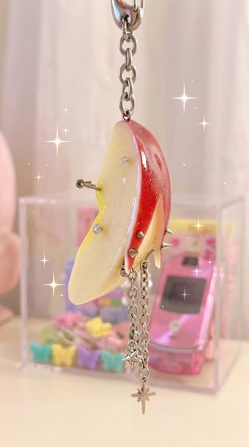 May include: A keychain featuring a realistic apple slice charm with a red skin and pale yellow flesh. The apple slice is decorated with small silver spikes, clear rhinestones, and a silver chain with star-shaped charms hanging from the bottom.