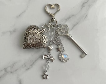 May include: A silver-toned keychain featuring a heart-shaped locket with intricate filigree, a skeleton key, and dangling charms including a crescent moon with a small iridescent heart, stars, a bow, and clear beads attached to a heart-shaped ring.