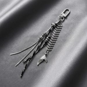 May include: A silver-toned keyring featuring a lobster clasp, a metallic fish skeleton charm, and multiple dangling strands. The strands include a smooth silver chain, a black textured cord, and a silver chain accented with small metallic beads.