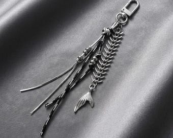 May include: A silver-toned keychain featuring a lobster clasp, a metallic fish skeleton charm, and multiple dangling strands. The strands include a smooth silver chain, a black textured cord, and a silver chain accented with small metallic beads.