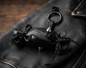 Könnte beinhalten: Ein schwarzes, bonbonförmiges Accessoire mit einem zentralen Beutel und drei silberfarbenen, konischen Nieten. Die Enden sind mit schwarzen Bändern mit kleinen weißen Punkten zu Schleifen gebunden. Ein schwarzer Metallring ist oben befestigt.