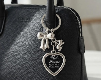 Könnte beinhalten: Eine schwarze Handtasche aus strukturiertem Leder mit einem silberfarbenen Schlüsselanhänger am Griff. Der Anhänger besteht aus einer Schleife, einem kleinen Hasen und einem herzförmigen Fotorahmen mit dem Text Photo Frame 2.9cm x 3.1cm.