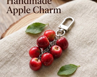 Könnte beinhalten: Ein Schlüsselanhänger mit einem Bündel aus sieben kleinen, glänzend roten Apfelnachbildungen an einem silberfarbenen Karabinerhaken. Der Text 'Handmade Apple Charm' ist im oberen Teil des Bildes zu sehen.