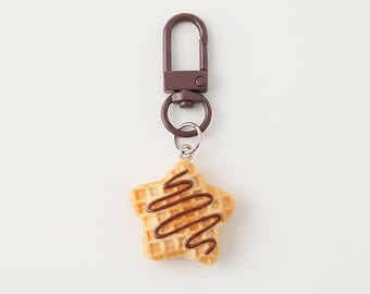 Könnte beinhalten: Ein sternförmiger Waffel-Schlüsselanhänger mit goldbrauner Gitterstruktur und einem dunkelbraunen Schokoladenguss. Der Anhänger ist mit einem kleinen silberfarbenen Ring an einem dunkelbraunen Metall-Karabinerhaken befestigt.