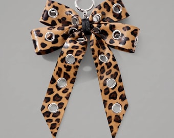 Könnte beinhalten: Ein Schleifen-Accessoire mit Leopardenmuster in Beige und Schwarz. Die Schleife besteht aus einem glatten Material und ist mit mehreren silberfarbenen Metallösen versehen, die über die Schlaufen und Enden verteilt sind. Ein silberner Karabinerhaken ist befestigt.