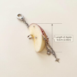 May include: A keychain featuring a slice of apple adorned with silver-toned metal piercings, including spikes, rings, and star charms on a chain. The apple slice measures 2.56 inches in length as indicated by the text in the image.