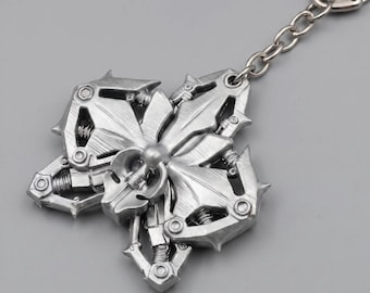Könnte beinhalten: Ein Schlüsselanhänger aus silberfarbenem Metall mit einem komplexen, mehrschichtigen mechanischen Design, scharfen, kantigen Formen und sichtbaren Spiralfedern. Das zentrale Ornament ist mit einer Metallkette und einem Schlüsselring verbunden.