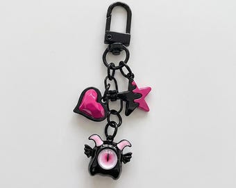 Könnte beinhalten: Ein Schlüsselanhänger aus schwarzem Metall mit Karabinerverschluss und einer Kette mit drei Anhängern: einem glänzenden pinken Herz, einem schwarz-pinken Stern und einer schwarzen Kreatur mit kleinen Flügeln, pinken Hörnern und einem Auge.