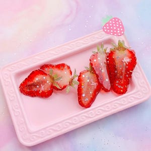 May include: A rectangular light pink tray with an embossed border holds four strawberry slices embedded with silver star-shaped glitter. A small pink strawberry-shaped charm with white polka dots is attached to the top right corner of the tray.
