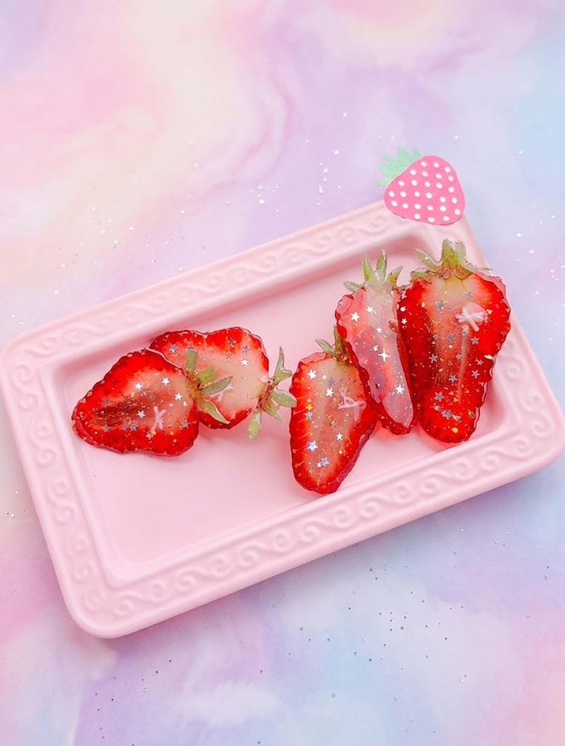 May include: A rectangular light pink tray with an embossed border holds four strawberry slices embedded with silver star-shaped glitter. A small pink strawberry-shaped charm with white polka dots is attached to the top right corner of the tray.