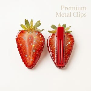 May include: Two hair clips shaped like strawberry slices with green leaves at the top. The red fruit surface is embedded with silver and gold star-shaped glitter. The back of one clip features a red metal alligator clip. Text reads Premium Metal Clips.