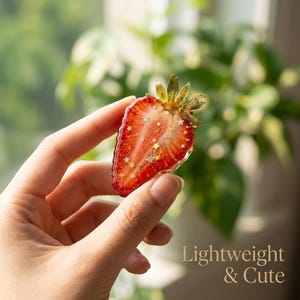 May include: A strawberry-shaped resin charm featuring a vibrant red interior with embedded gold and silver star-shaped glitter, topped with a green stem. The piece has a glossy, translucent finish and is held up to show its size and detail.