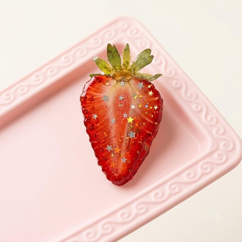 May include: A resin strawberry slice ornament featuring a vibrant red interior with embedded silver and gold star-shaped glitter and a green leafy stem at the top, resting on a light pink rectangular tray with a decorative embossed border.