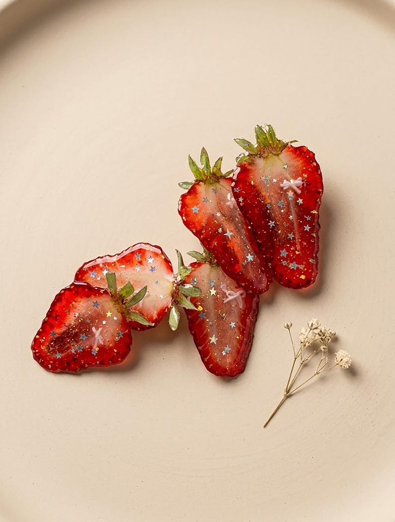 May include: Three strawberry-shaped resin ornaments featuring a translucent red finish, green leafy tops, and embedded silver star-shaped glitter. Each piece displays a glossy texture with small white accents, accompanied by a single dried flower sprig.