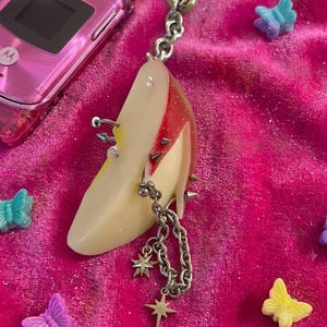 Könnte beinhalten: Ein Schlüsselanhänger mit einem realistischen Apfelschnitz-Anhänger, verziert mit silberfarbenen Metallspikes, Nieten und einer hängenden Kette mit zwei vierzackigen Sternanhängern. Der Apfelschnitz hat eine rote Schale und helles Fruchtfleisch.