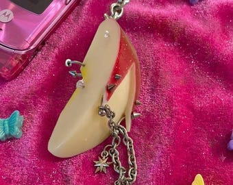 Könnte beinhalten: Ein Schlüsselanhänger mit einem realistischen Apfelschnitz-Anhänger, verziert mit silberfarbenen Metallspikes, Nieten und einer hängenden Kette mit zwei vierzackigen Sternanhängern. Der Apfelschnitz hat eine rote Schale und helles Fruchtfleisch.