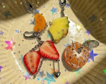 Könnte beinhalten: Ein Set aus fruchtförmigen Schlüsselanhängern mit einer leuchtend orangefarbenen Zitrusscheibe, einem gelben Ananasstück, zwei roten Erdbeerscheiben und einer ganzen Orange, verziert mit kleinen silbernen Nieten und silbernen Karabinerhaken.